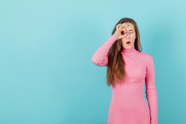 La fille blonde fait un cercle avec ses doigts autour de son oeil sur le fond bleu