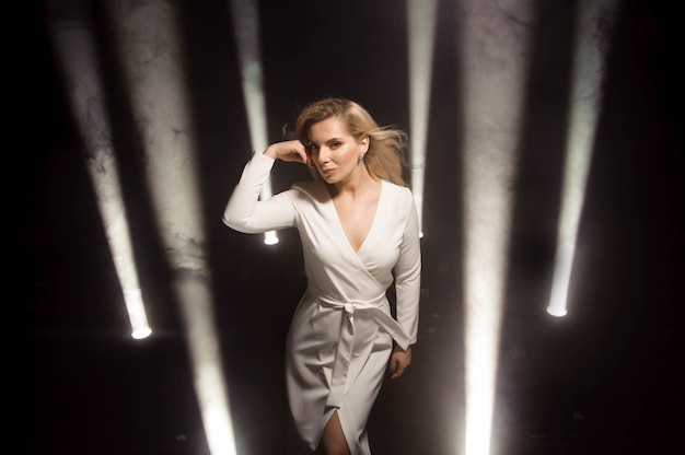 Fille blonde fashion avec de longs et brillants cheveux bouclés. Beau modèle en robe blanche sur la scène avec des lumières.