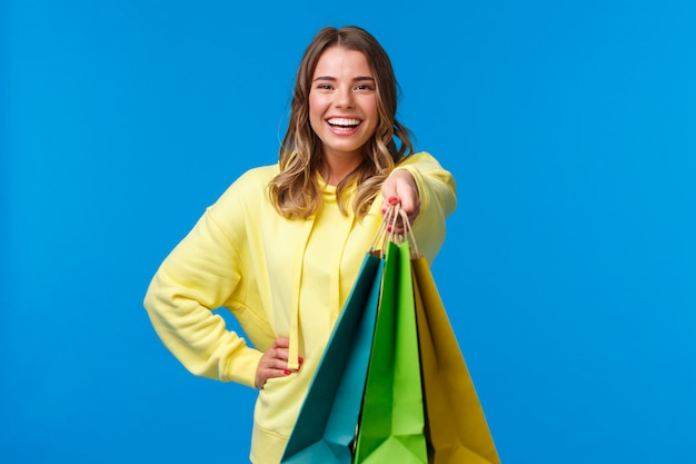 Fille blonde gaie vous donnant des sacs à provisions et souriant heureux, achetant des cadeaux pour les vacances en famille, achetant beaucoup de personnel en magasin et demandant de porter ses colis, stand