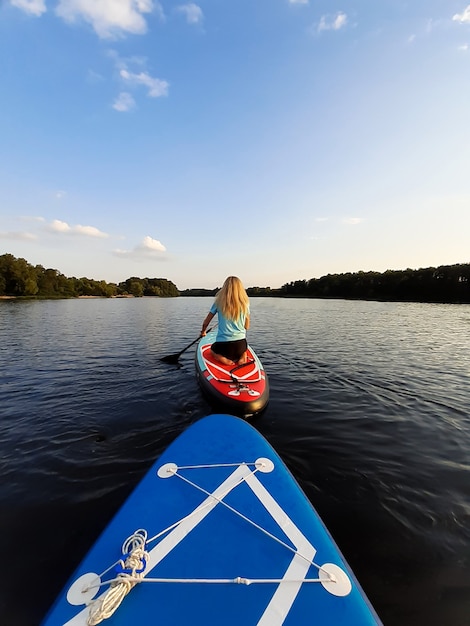 Une fille blonde à genoux flotte sur une planche à pagaie SUP, s'aidant d'une rame. La deuxième planche flotte derrière. Vue arrière