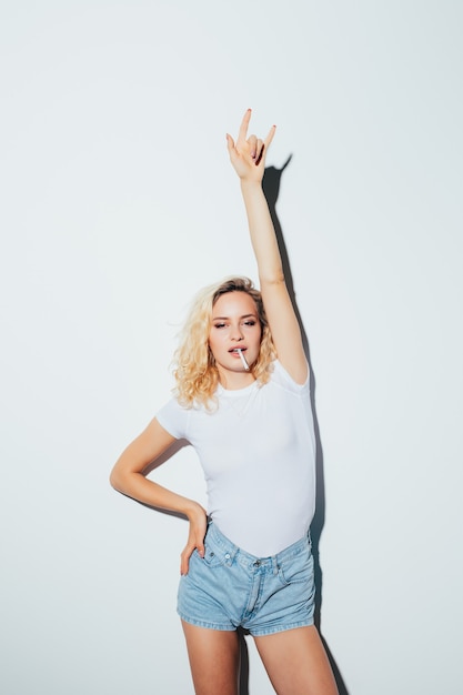 Fille blonde hipster en t-shirt blanc cigarette et donnant le signe Rock and Roll isolé sur mur blanc