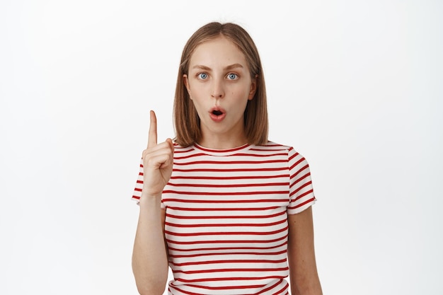 Fille blonde impressionnée pointant le doigt vers le haut, haletant et regardant la caméra étonnée, portant un t-shirt, montrant une publicité, une bannière sur un espace vide, fond blanc. Copier l'espace