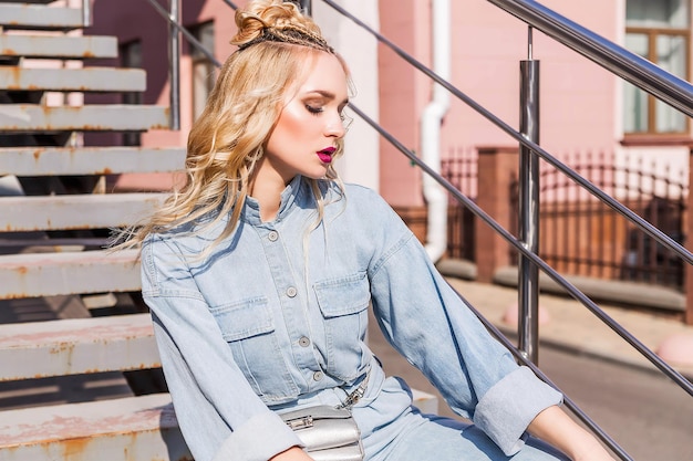 Une fille blonde avec un maquillage lumineux et une coiffure en salopette en jean est assise sur les escaliers de la rue