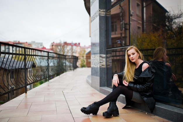 Fille blonde à la mode en long manteau de cuir noir posé contre une grande fenêtre du bâtiment