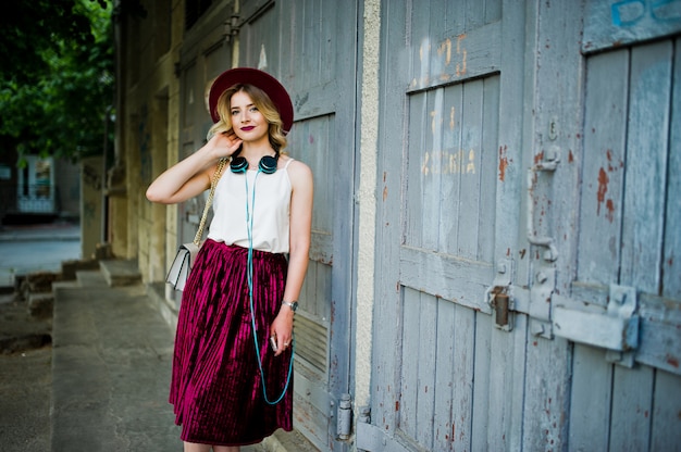 Fille blonde modèle à la mode et belle en élégante jupe en velours rouge velours, chemisier blanc et un chapeau, posé avec téléphone et écouteurs.
