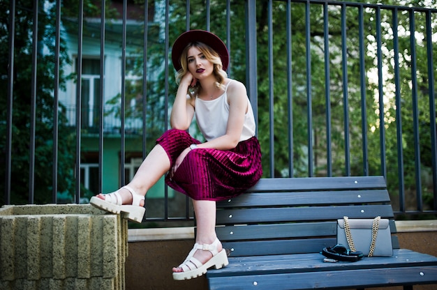 Fille blonde modèle à la mode et belle en élégante jupe en velours rouge velours, chemisier blanc, chapeau et sac à main, posée sur un banc.