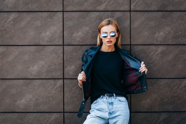 Fille blonde portant un t-shirt noir, des lunettes et une veste en cuir posant contre stree