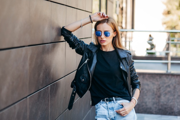 Fille blonde portant un t-shirt noir, des lunettes et une veste en cuir posant contre stree