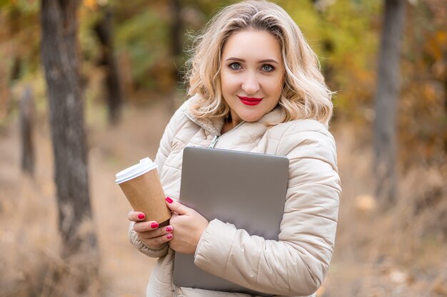 Une fille blonde qui est dans un parc d'automne a saisi un ordinateur portable et une tasse de café