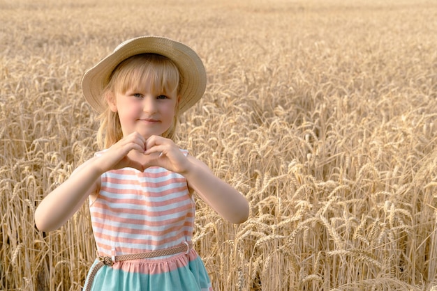 Une fille blonde regarde la caméra et montre le cœur de ses mains dans le champ de blé