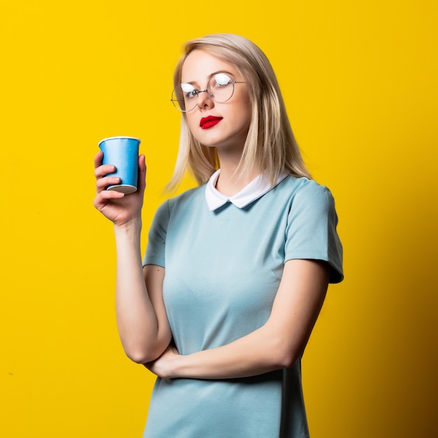 Fille blonde en robe bleue et verres et tasse de papier sur fond jaune