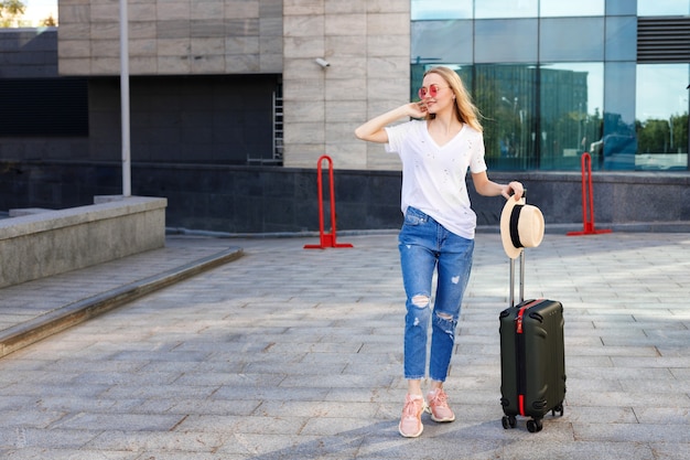 Fille blonde avec sac de voyage et chapeau de paille attendant l'été de bus à l'aéroport