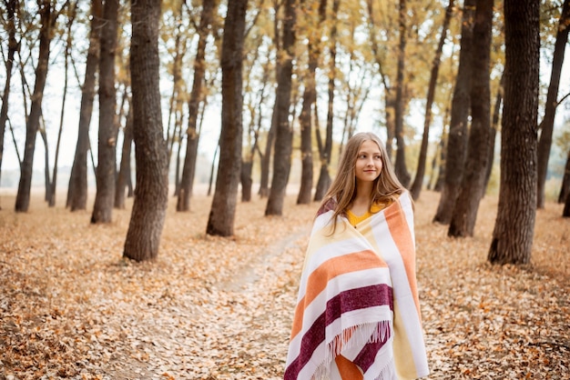 Une fille blonde se couvre d'une couverture pour se réchauffer, se promenant dans le parc en automne. Temps frais, temps libre sur la nature, bonne humeur.