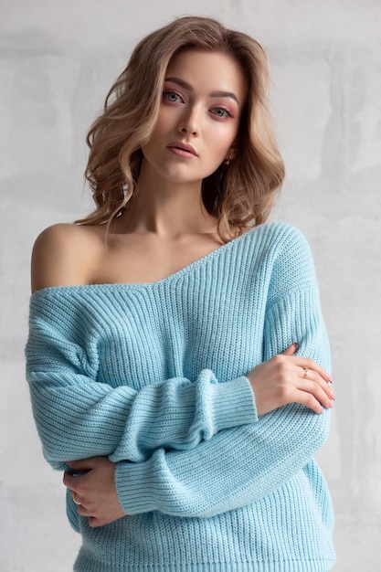 Fille blonde sexy dans un pull en tricot bleu sur un fond de mur blanc