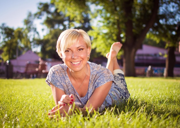 Une fille blonde souriante avec une coupe de cheveux courte est allongée dans un parc de la ville sur l'herbe verte Une femme se repose dans un parc et regarde la caméra