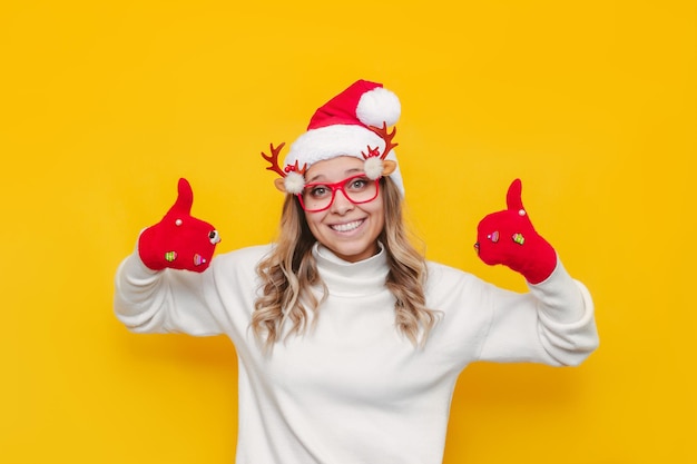 Une fille blonde souriante dans des mitaines rouges de bonnet de Noel de Noël montre le pouce vers le haut comme un geste avec ses mains