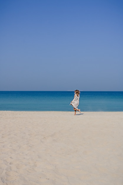 Une fille blonde souriante dans une robe maxi en lin avec des cheveux flottants sautant et dansant sur une plage de sable