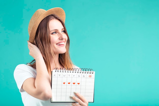 Une fille blonde souriante porte un t-shirt blanc et un chapeau tenant un calendrier de vacances sur fond bleu concept de destination estivale