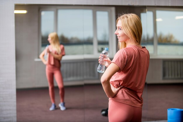 Une fille blonde souriante tient une bouteille d'eau à la main dans la salle de sport