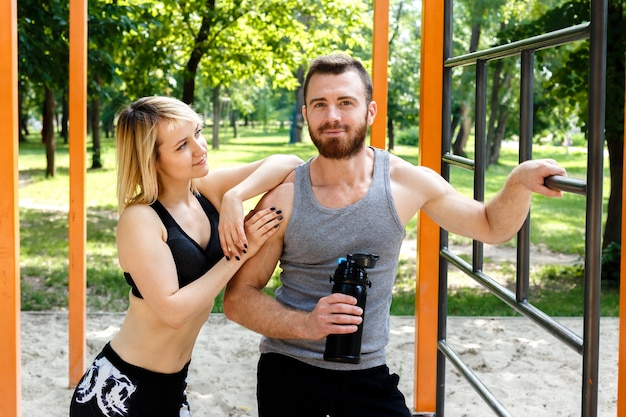 Fille blonde sportive et homme barbu se reposer après une formation d'entraînement dans un parc en plein air. Homme tenant une bouteille noire avec de l'eau.