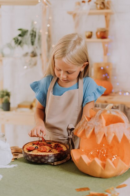 fille blonde en tablier dans la cuisine décorée de citrouilles pour Halloween prépare une tarte focaccia