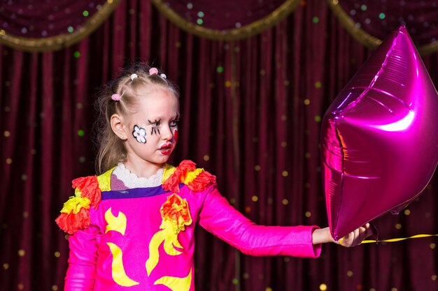 Fille blonde talentueuse jouant avec un diadème coloré et des fleurs peintes sur le visage et cousues sur un costume de théâtre rose vif