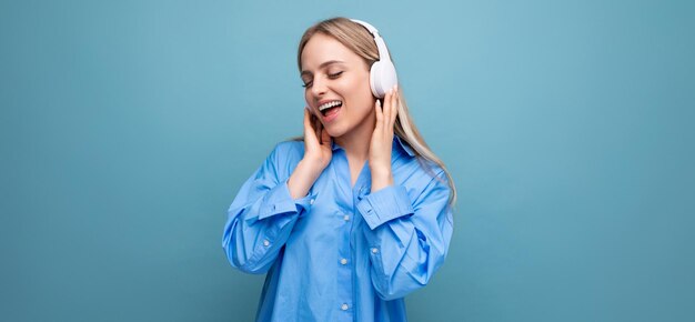 Une fille blonde timide dans une chemise décontractée écoute des chansons dans de gros écouteurs et chante sur un bleu