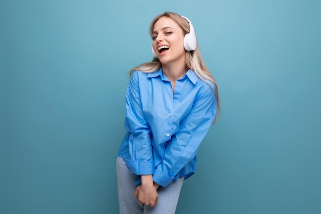 Une fille blonde timide dans une chemise décontractée écoute des chansons dans de gros écouteurs et chante sur un bleu