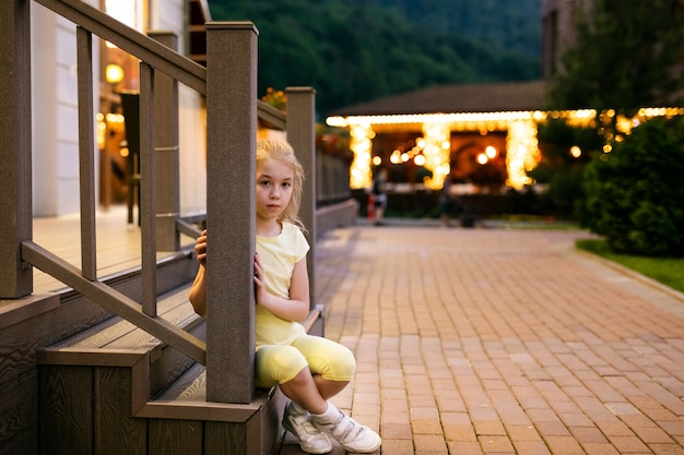 Fille blonde triste de 9 ans en habits jaunes assis sur les traces de bois du restaurant