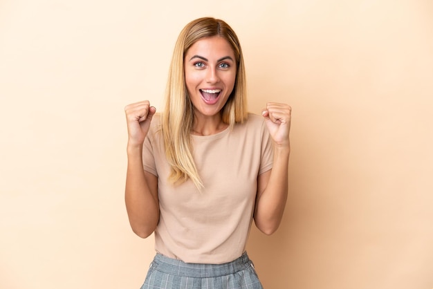 Fille blonde uruguayenne isolée sur fond beige célébrant une victoire en position de vainqueur