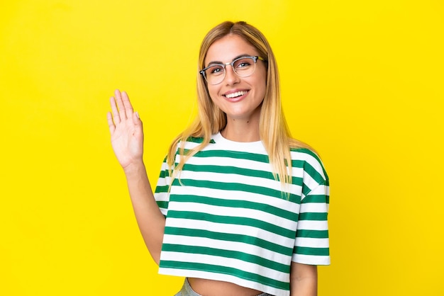 Fille blonde uruguayenne isolée sur fond jaune saluant avec la main avec une expression heureuse