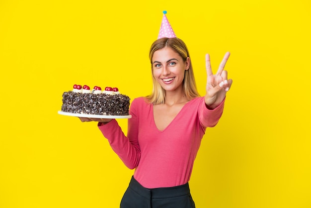 Fille blonde uruguayenne tenant un gâteau d'anniversaire isolé sur fond jaune souriant et montrant le signe de la victoire