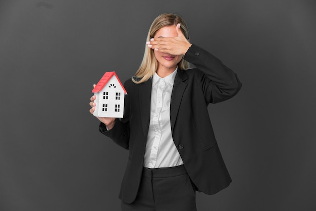 Fille blonde uruguayenne tenant un jouet de maison isolé sur fond noir couvrant les yeux par les mains Je ne veux pas voir quelque chose