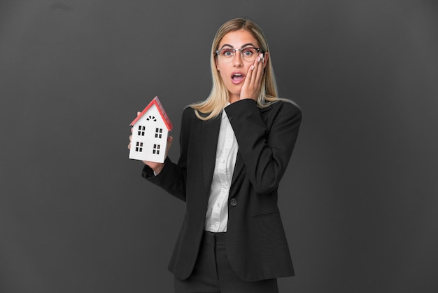 Fille blonde uruguayenne tenant un jouet de maison isolé sur fond noir avec une expression faciale surprise et choquée