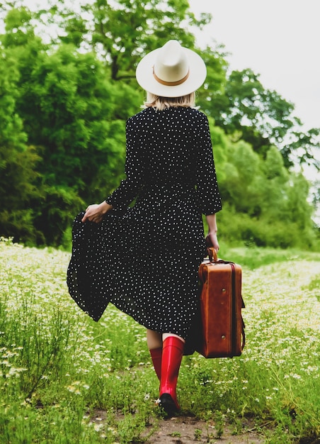 Fille blonde avec valise et chapeau dans le champ de fleurs de camomille en été
