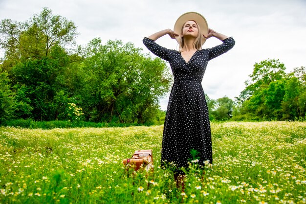 Fille blonde avec valise et chapeau dans le champ de fleurs de camomille en été