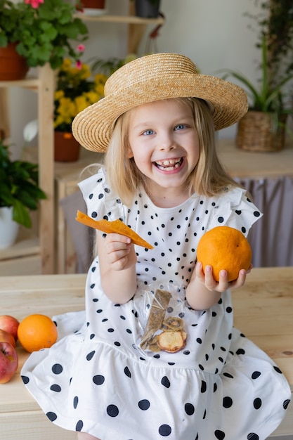 fille blonde vêtue d'une robe blanche avec des pois noirs tient une orange pelée dans ses mains