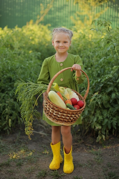 fille blonde vêtue d'une robe verte et de bottes jaunes se dresse dans le jardin et détient des légumes cueillis