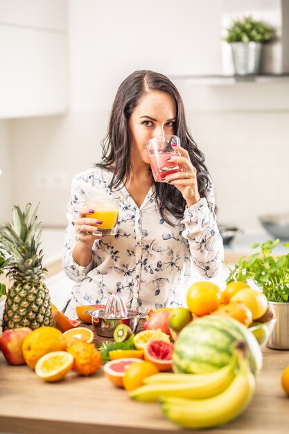 Une fille boit du jus de fruits frais à la maison avec d'autres types de fruits sur le bureau de la cuisine devant elle.