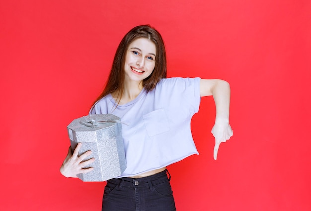 Fille avec une boîte-cadeau en argent remarquant et invitant la personne devant la recevoir.