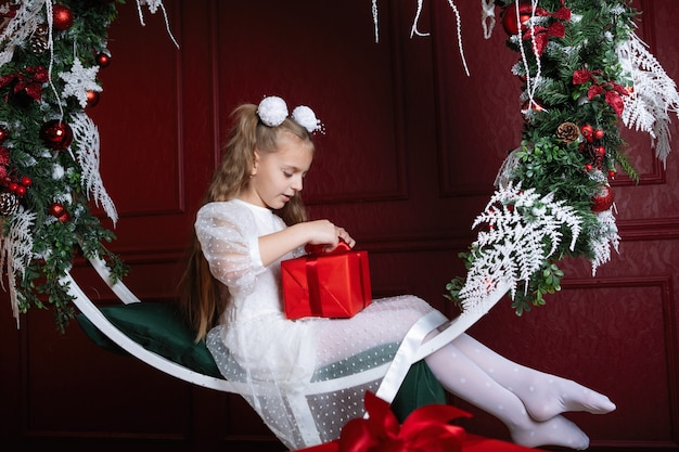 Fille avec la boîte rouge. Cadeau de Noël. L'adolescent est assis sur la balançoire en arc avec des couronnes de décoration du Nouvel An, des branches d'épinette, des boules et des guirlandes.