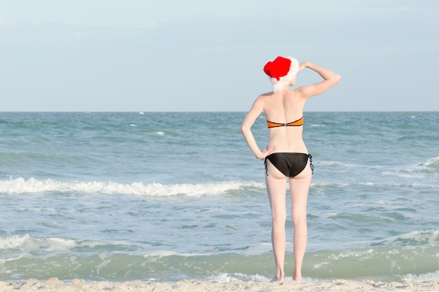 Fille en bonnet de Noel et maillot de bain se penche sur la distance. Bord de mer.