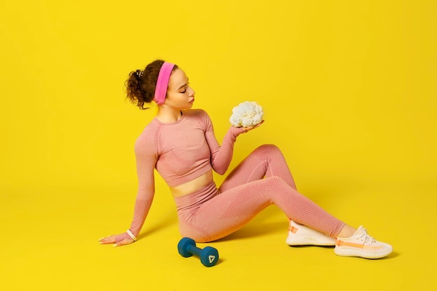 Une fille bouclée athlétique en vêtements de sport roses serrés est assise sur le sol et tient du chou-fleur dans ses mains