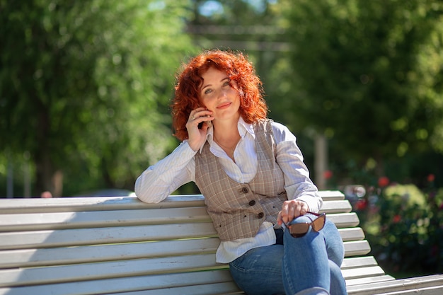 Fille bouclée aux cheveux roux dans une chemise et un jean est assise sur un banc dans le parc et parle au téléphone