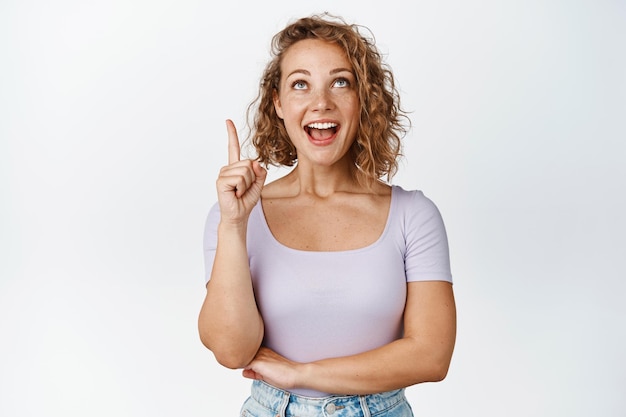 Fille bouclée blonde excitée, pointant le doigt et levant les yeux avec une expression de visage souriante fascinée, debout en t-shirt sur fond blanc