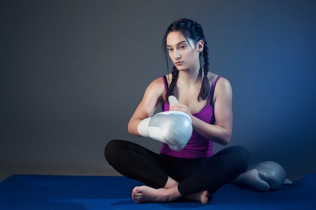 Une fille boxeuse décolle enfiler des gants de boxe tout en restant assise sur le tapis