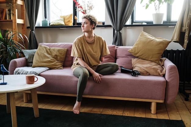 Fille avec un bras amputé assis sur un canapé dans le salon avec une prothèse elle a l'air pensive