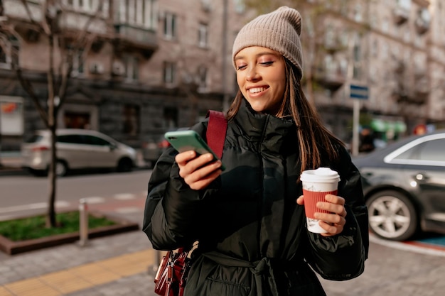 une fille brillante avec un magnifique sourire vêtu d'une casquette beige et d'une veste sombre discute en ligne avec des amis et boit du café en plein air par une chaude journée ensoleillée dans le centre-ville