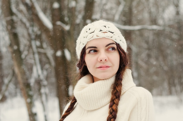 Fille brune aux cheveux longs marchant dans un parc d'hiver et jouant avec la neige, vêtu d'un pull blanc et d'un chapeau blanc fait main