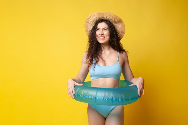 fille brune élancée et bouclée en maillot de bain bleu en été avec un anneau de natation gonflable posant